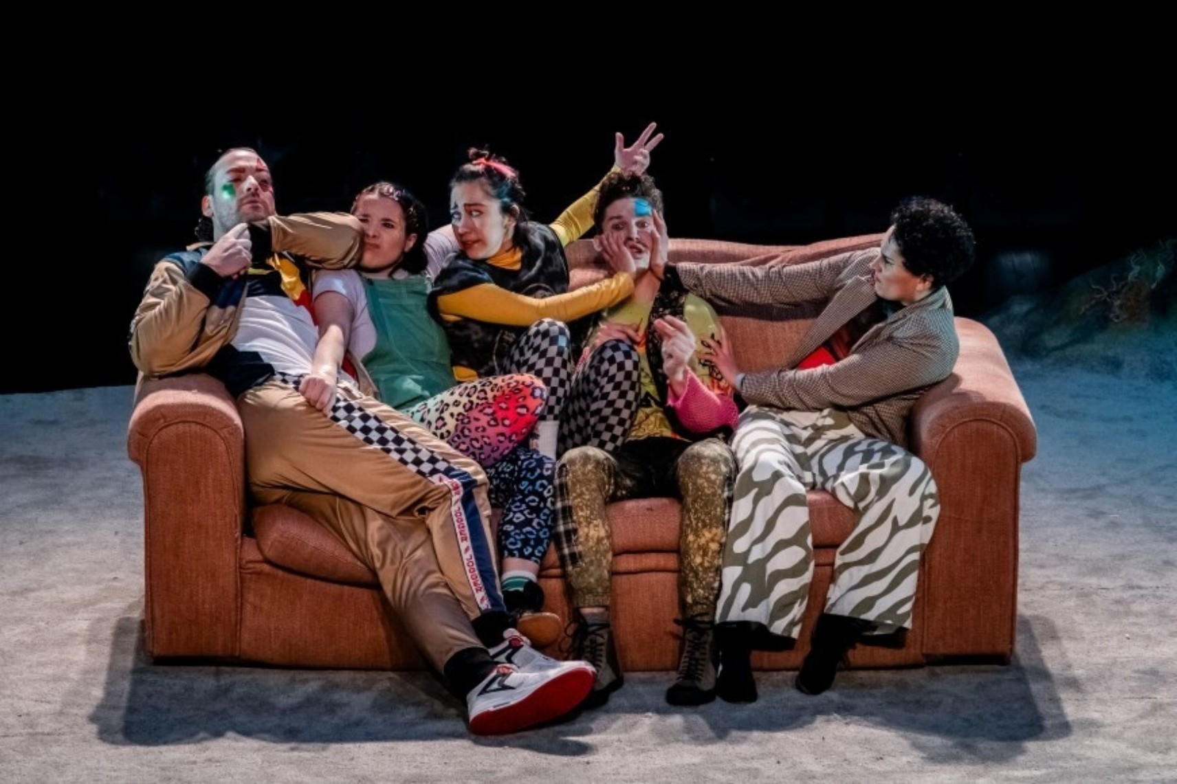 Cinq personnages s'entassent sur un divan, pressant leurs mains dans les visages des autres. Tous les personnages portent des vêtements colorés et à motifs variés, et ont des visages expressifs.