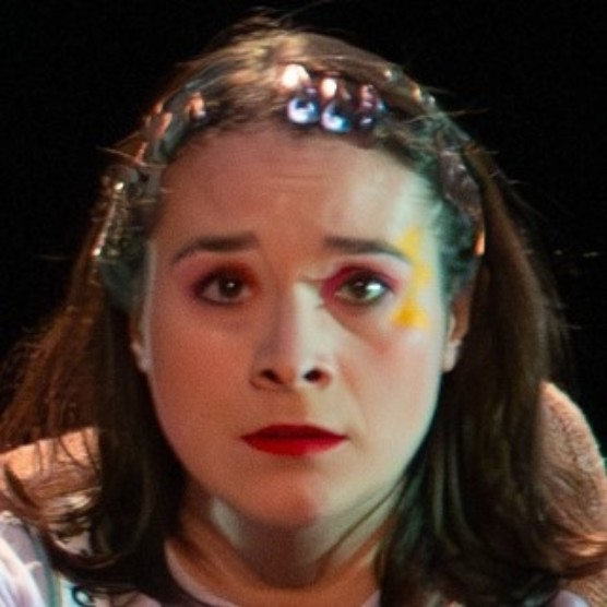La comédienne a des barrettes métalliques dans les cheveux tout le tour du front. Elle a du rouge à lèvres rouge et un maquillage coloré. Elle regarde devant elle les sourcils levés vers le centre.