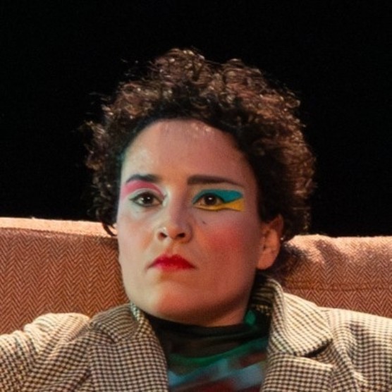 La comédienne est adossée sur un divan et fait la moue. Elle porte un veston brun, du rouge à lèvres rouge et un maquillage coloré.