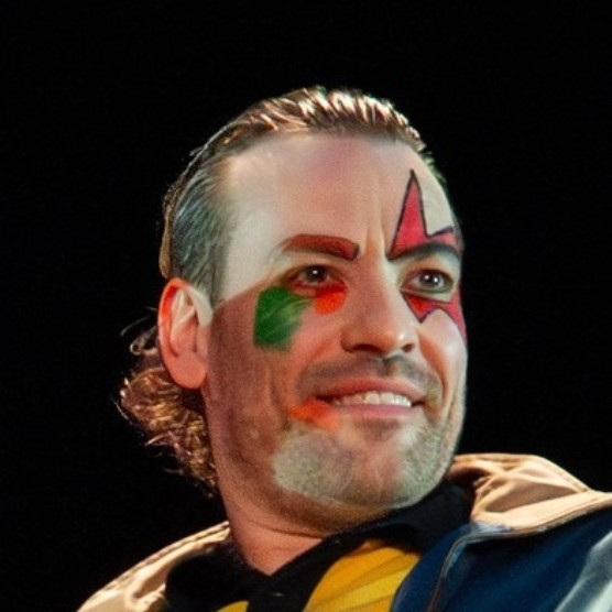 Le comédien a les cheveux coiffés vers l'arrière et un maquillage théâtral coloré. Son visage est tournée vers la droite et il regarde devant lui en souriant.