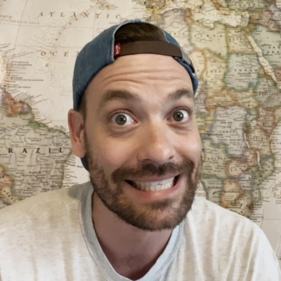 Photo de Marc Andre, un homme au teint clair, aux yeux et aux cheveux foncés, portant une barbe courte. Il porte un chandail gris et regarde la caméra avec les yeux écarquillés et un grand sourire. Derrière lui, une mappemonde.