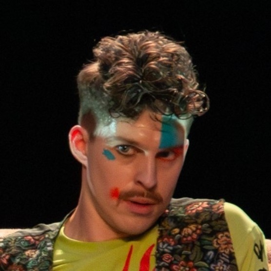 Le comédien porte un t-shirt vert lime, un boléro fleuri et un maquillage théâtral coloré. Son visage est légèrement penché et il regarde vers la gauche.