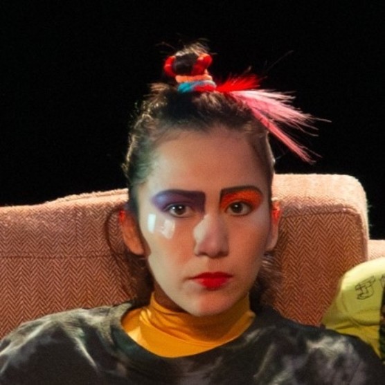 La comédienne est adossée à un divan. Ses cheveux sont attachés en toque avec des accessoires colorés. Elle porte un chandail tie dye foncé, du rouge à lèvres rouge, un maquillage coloré et elle regarde droit devant elle.