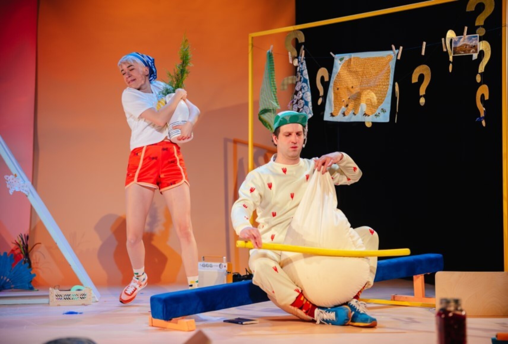 Soeur danse avec une plante dans un pot et Frère semble jouer du violoncelle avec un gros coussin, beige comme son costume. Les deux sont dans un décor rempli d'objets, devant un fond orange et noir.