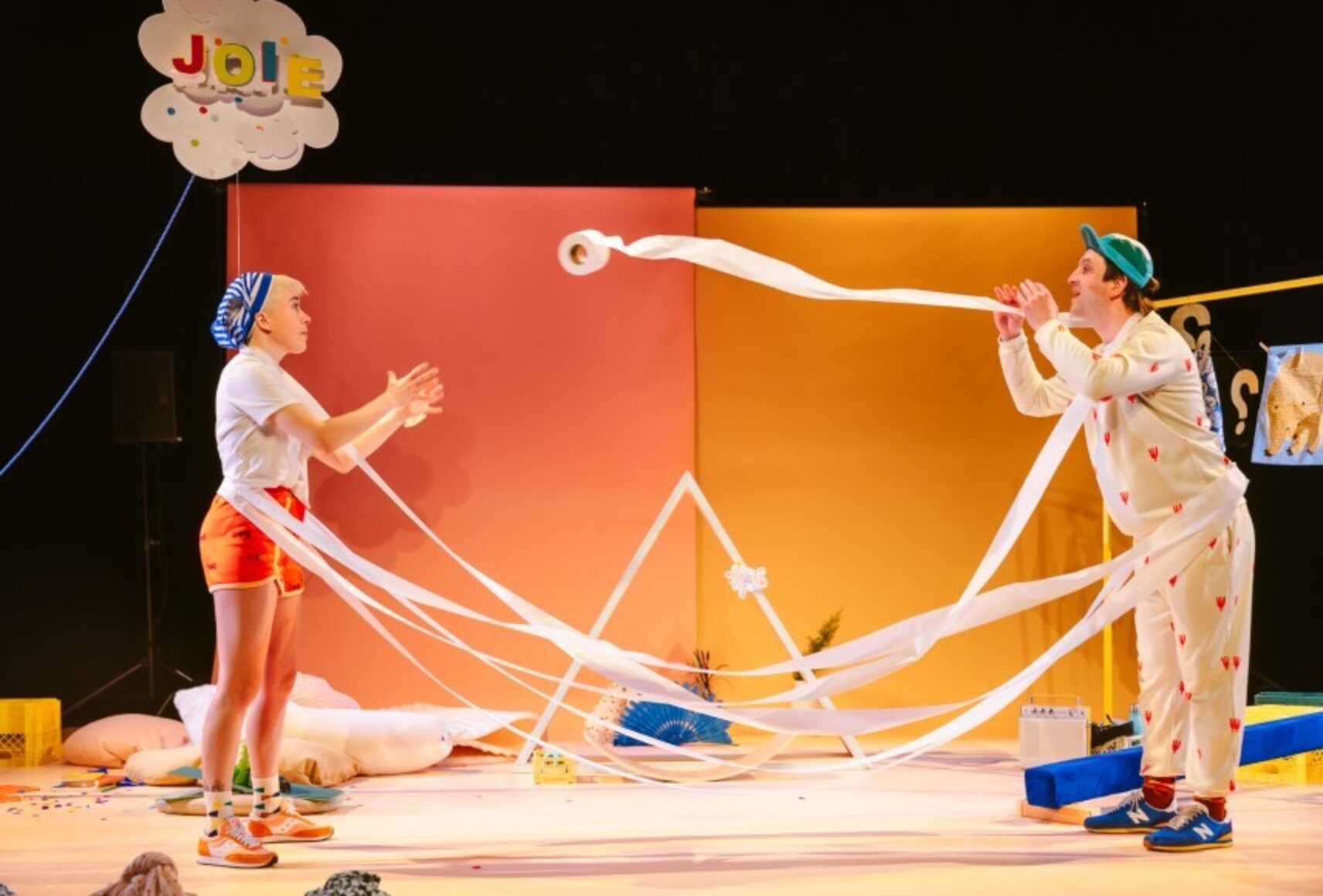 Deux personnages, Soeur avec un bandana et Frère avec une casquette, se lancent des rouleaux de papier de toilette devant un décor aux couleurs douces, jaune et rose saumon. En haut à gauche, le mot "Joie" écrit dans un nuage.