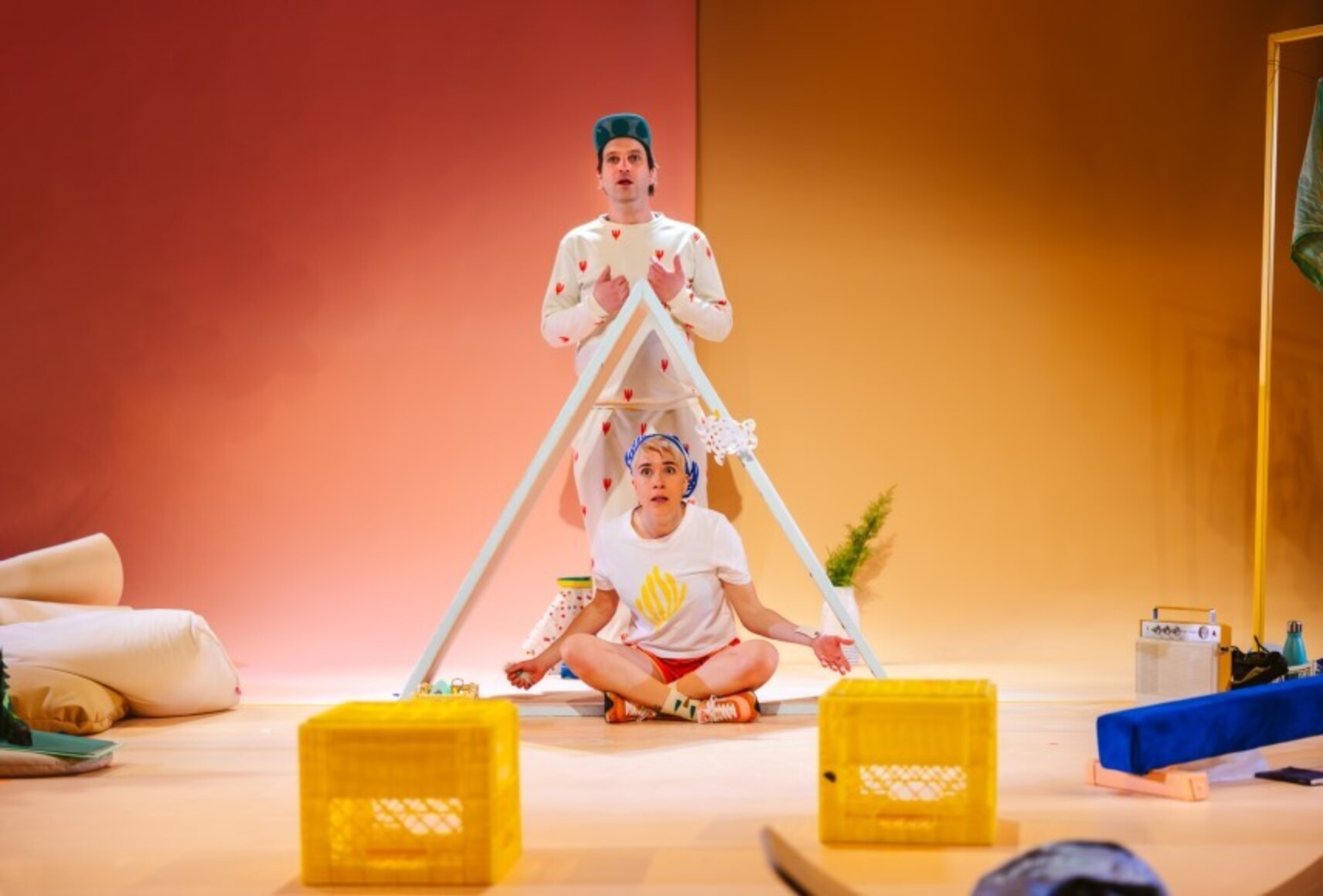 Frère est debout derrière Soeur, assise en tailleur sous un élément de décor en triangle rappelant le toit d'une maison. Des accessoires jaunes et bleus jonchent le sol. Le mur du fond est rose saumon à gauche et orange à droite.