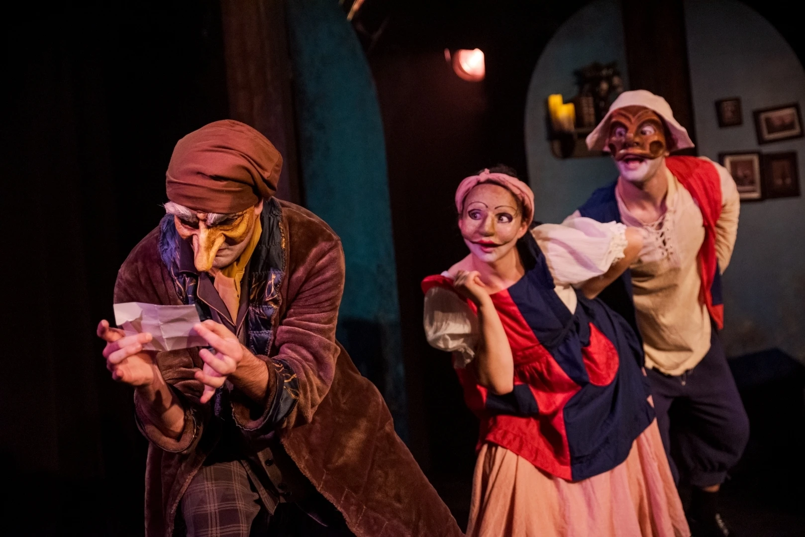 Photo de 3 acteurs masqués : un homme portant un masque de vieillard qui lit un petit papier, une femme avec un masque beige et une robe rouge et bleue qui semble curieuse, et un homme avec un masque plus large et brun, avec un large sourire.
