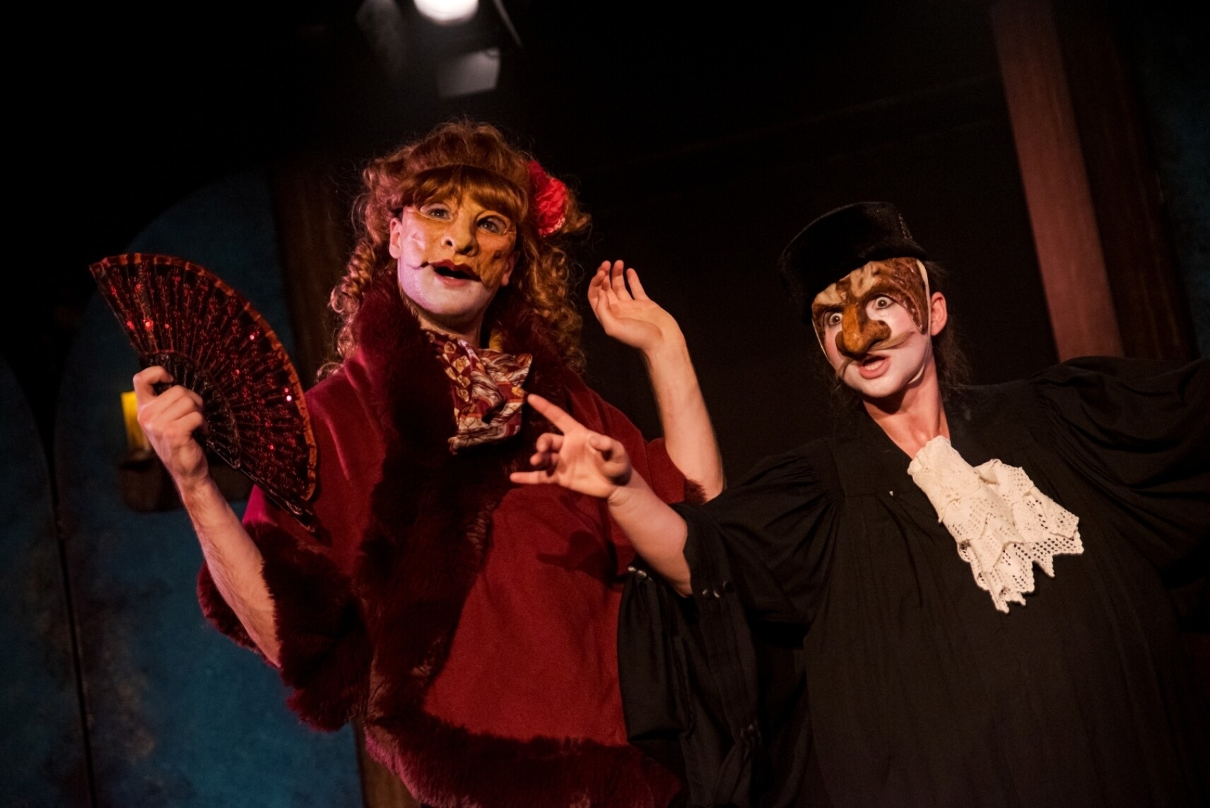 2 comédiens portent des masques de théâtre expressifs. La personne de gauche porte une perruque rousse bouclée, un costume rouge et un éventail décoratif. La personne de droite porte une grande fraise blanche en dentelle et un chapeau sombre..