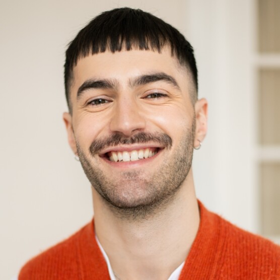 on voit un jeune homme souriant. Il a les cheveux noirs courts avec une frange droite, une moustache et une barbe légère. Il porte un t-shirt blanc sous un gilet sans manches en laine orange vif. Son expression est ouverte et joyeuse.