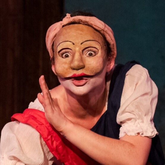 Sur cette photo, une comédienne masquée porte une jupe longue rosée avec un tablier rouge et bleu, une chemise blanche et un foulard rose sur la tête. Accroupie, un bras tendu et l’autre plié, elle adopte une posture expressive et comique.