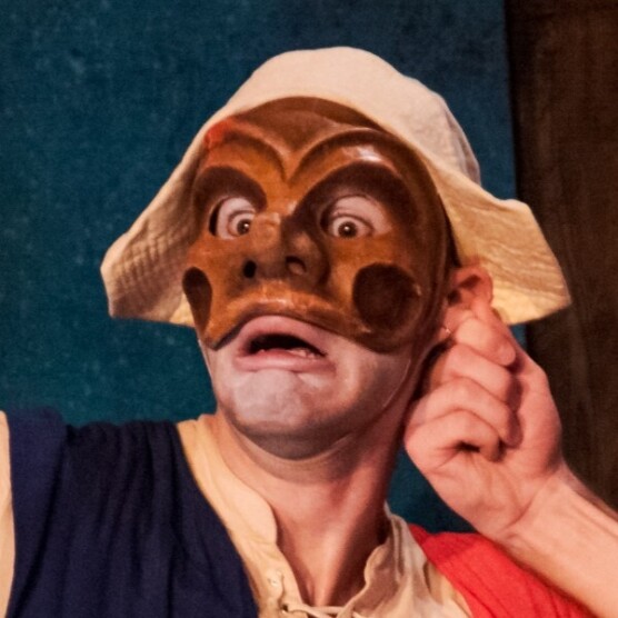 Sur cette photo, le personnage d’Arlequin, avec son masque brun au grand nez arrondi et ses yeux écarquillés, affiche une expression de frayeur comique, comme surpris ou pris au dépourvu.