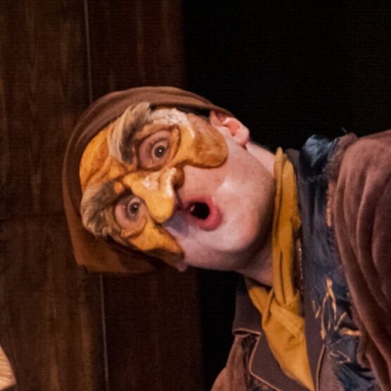 on voit un vieillard, portant un masque brun et jaune, qui a des sourcils froncés et de grands yeux ronds qui lui donnent un air étonné. Sa bouche entrouverte accentue l’expression de surprise comique, typique de la commedia dell’arte.