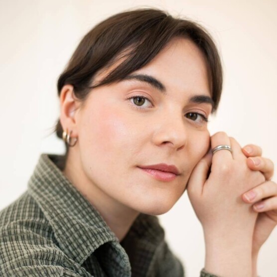 On voit une femme aux cheveux bruns courts et sourcils épais qui porte de petites boucles d’oreilles et une bague. Son teint est clair et elle porte une chemise à motif discret. Elle pose la tête légèrement inclinée, les mains jointes près du visage.