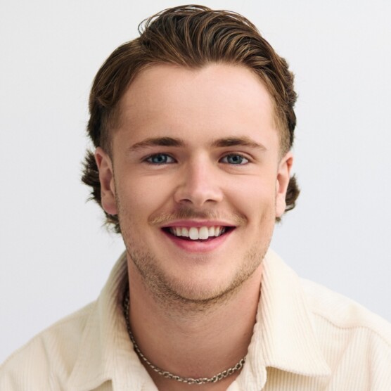 on voit un homme souriant, au visage jeune et lumineux. Il a les cheveux châtains coiffés vers l’arrière, les yeux bleus et une fine moustache. Il porte une chemise beige claire à gros boutons et un collier en chaîne métallique.
