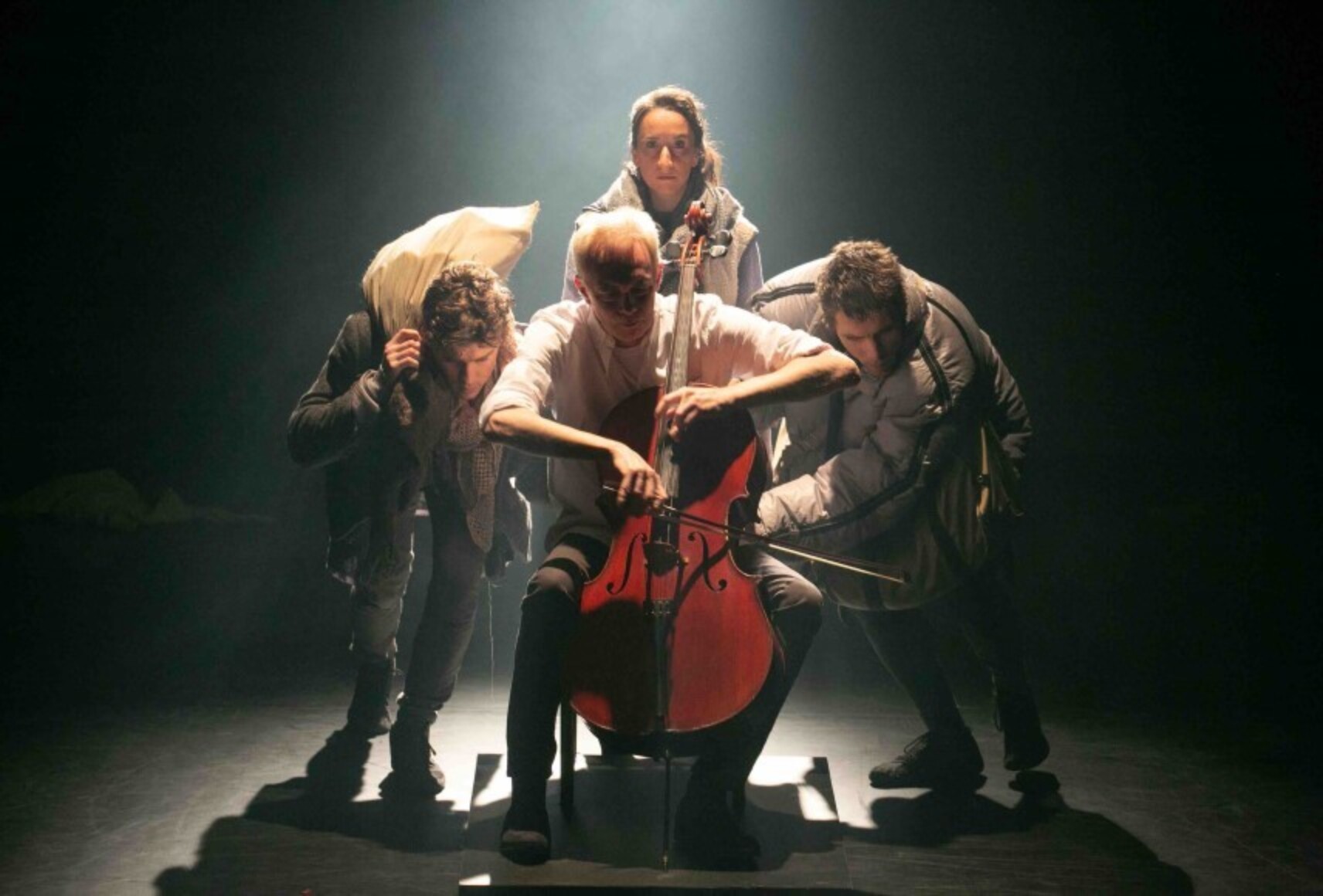 Photo des trois interprètes costumés, entourant une quatrième personne au centre, qui joue du violoncelle, sous une douche de lumière blanche.