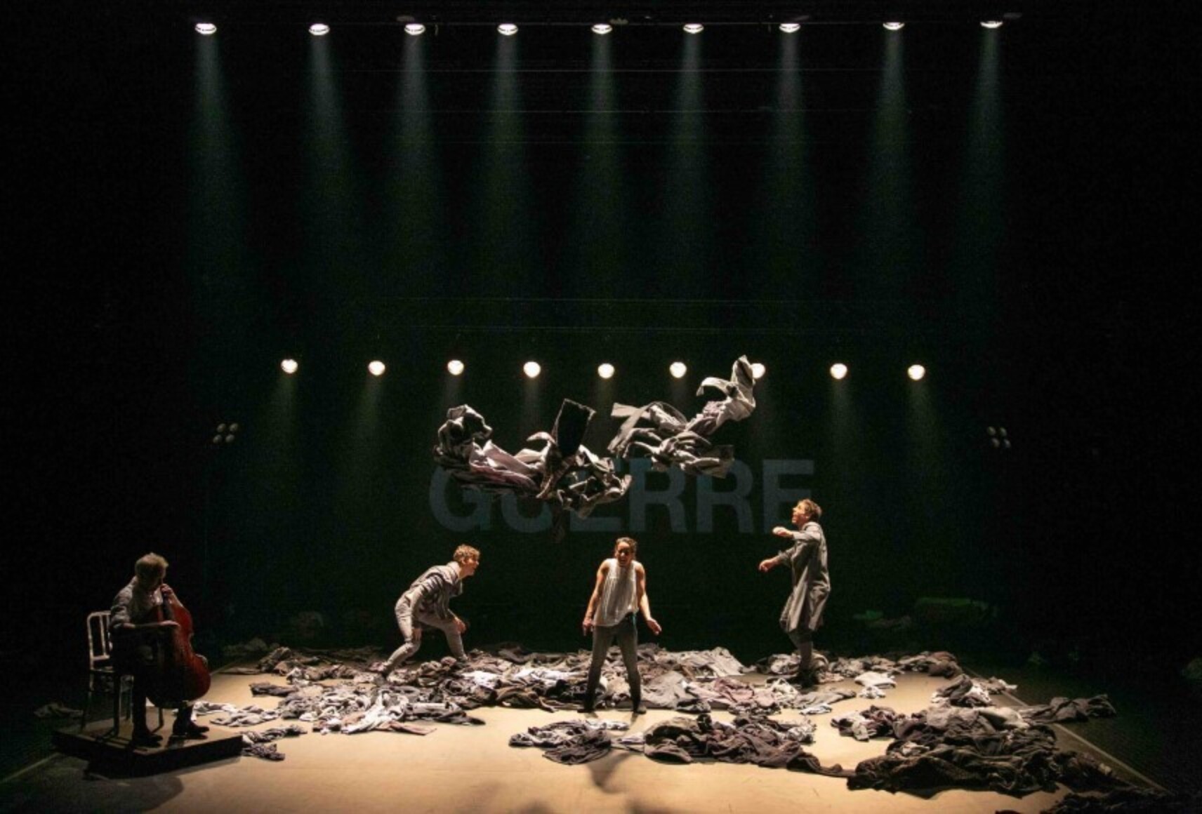 Photo en clair-obscur des quatre artistes devant la projection du mot "guerre". La scène est remplie d'objets texturés dont certains sont suspendus au-dessus de la scène.