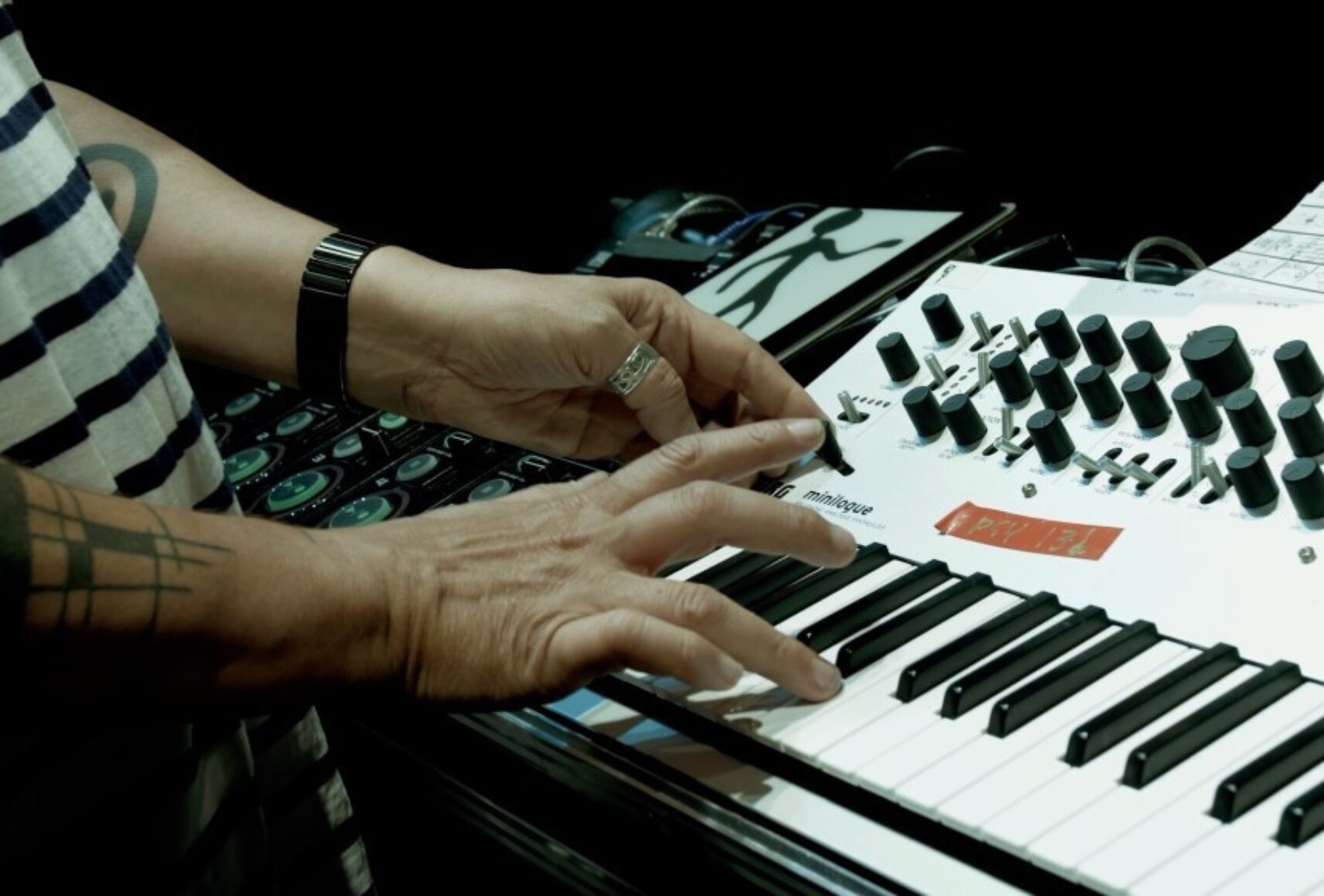 Gros plan sur les mains de Nicolas sur le clavier du synthétiseur. Son chandail rayé noir et blanc fait un rappel avec les touches noires et blanches.