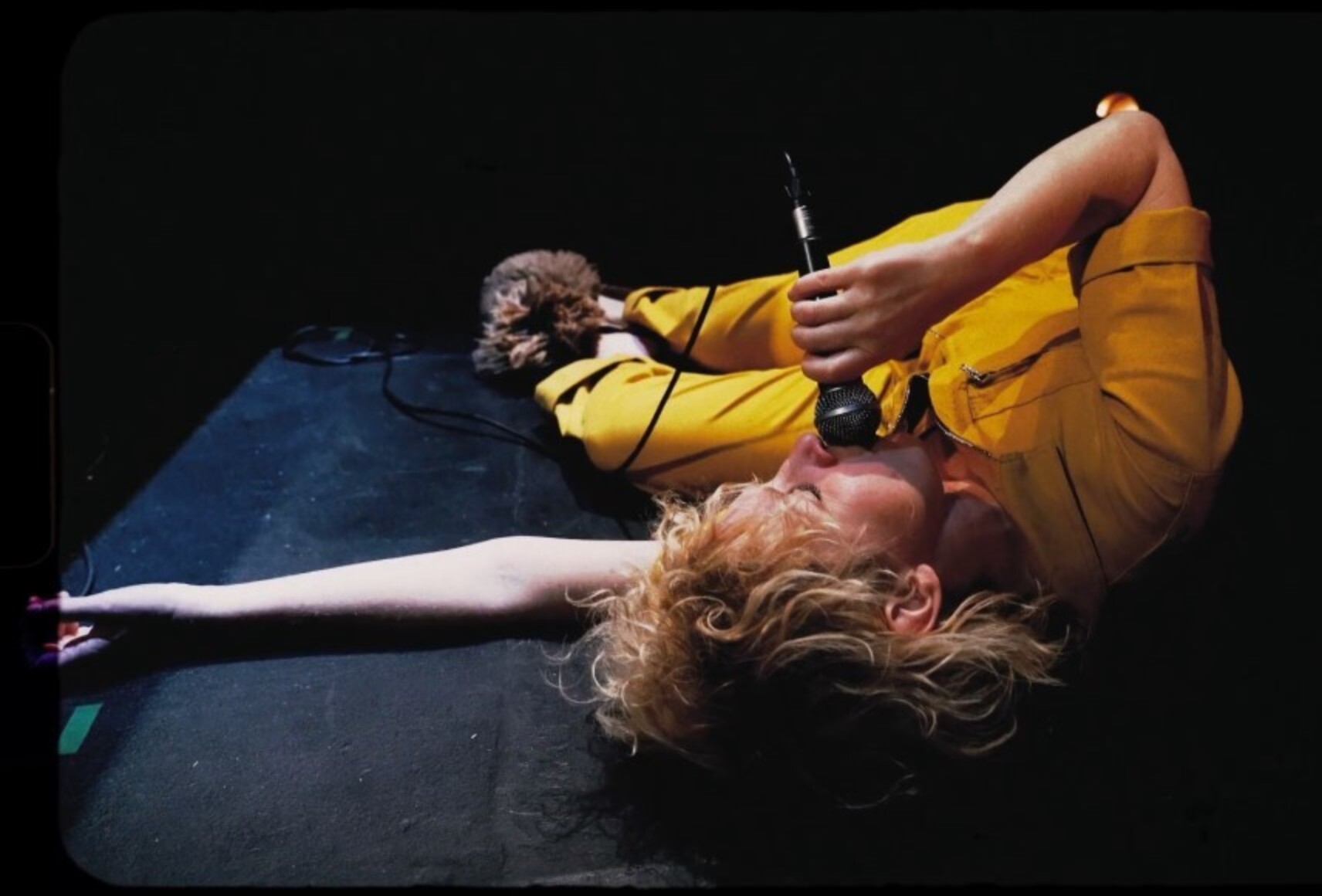 Photo de la comédienne Karine qui chante couchée au sol. Elle porte un habit jaune et tient le micro avec fil collé à sa bouche. Elle est éclairée par une douche de lumière blanche.