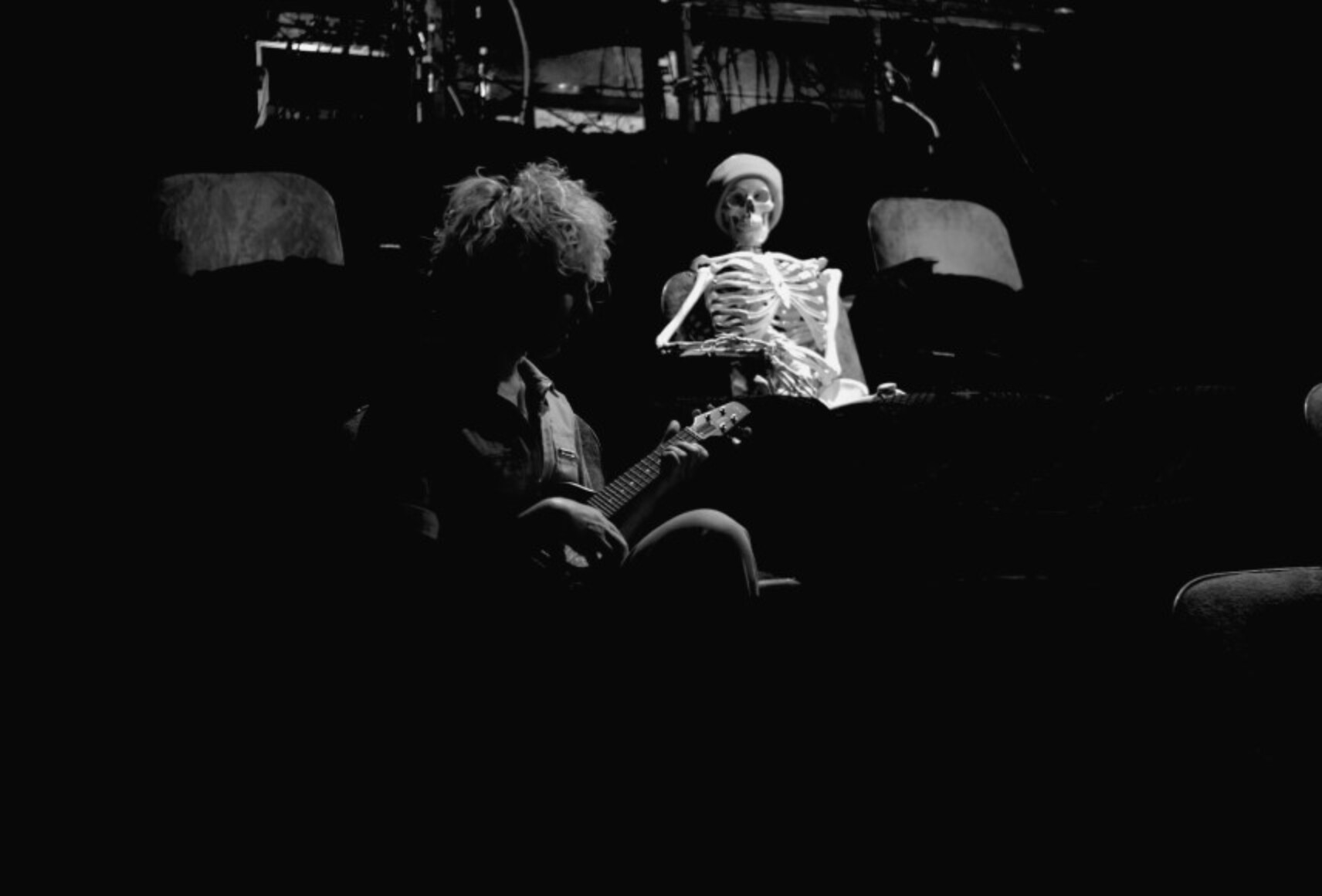 Photo en noir et blanc du psy-squelette regardant Karine qui joue du ukulele. Le squelette d'anatomie porte une tuque et est assis sur un banc de salle de spectacle.