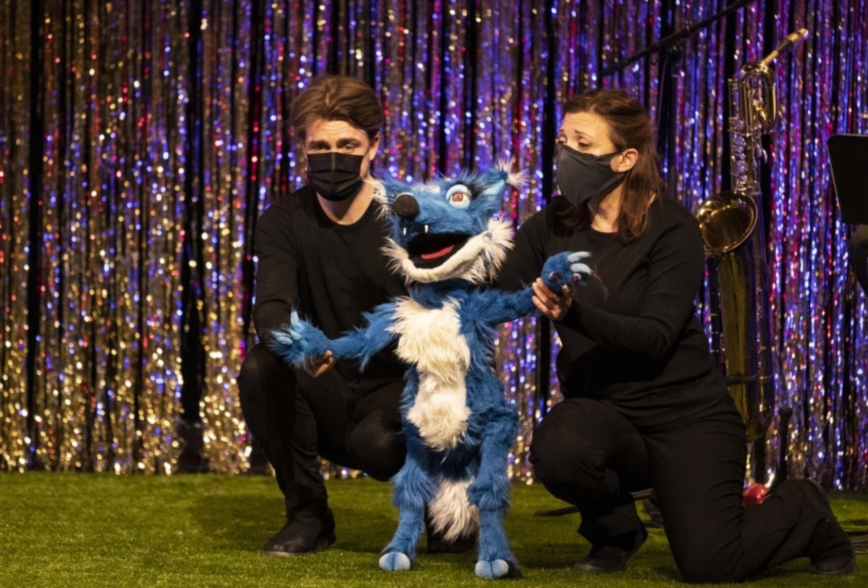 Photo de d'un homme et une femme manipulant une marionnette de renard au pelage bleu et blanc. Ils sont accroupis à ses côtés. Derrière eux, un rideau à franges scintillant.