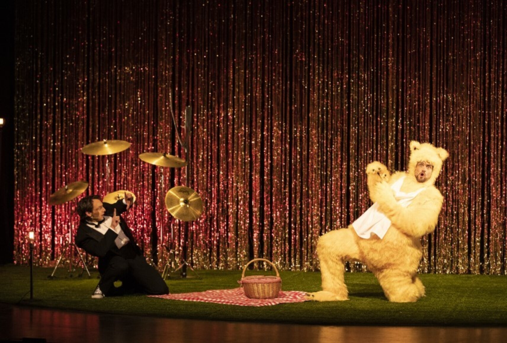 Un homme en tuxedo noir et blanc et un comédien en costume d'ours au pelage beige sont sur un tapis vert, une nappe et un panier de pique-nique entre eux. L'homme mime de prendre l'ours en photo. Derrière, des cymbales et un rideau à franges scintillant.