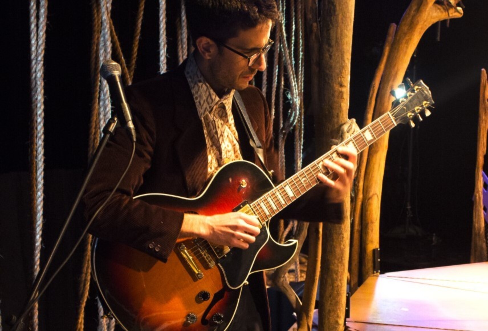 Photo d'un musicien jouant de la guitare. Il a les cheveux bruns, des lunettes et porte une veste noire. Un micro et un décor de cordage et de pièces de bois sont aussi visibles.