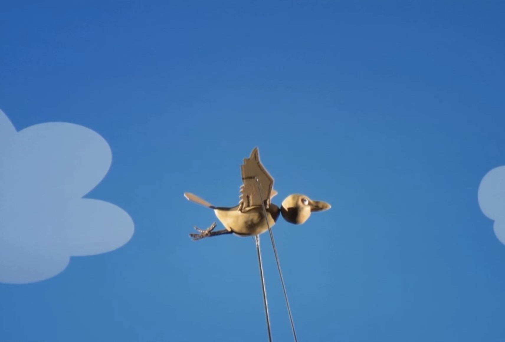 La marionnette de Coucou a déployé ses ailes et vole dans un ciel bleu au niveau des nuages.