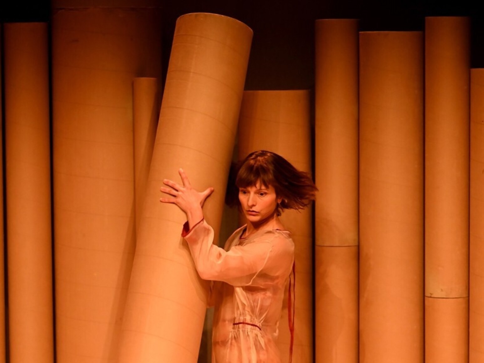 Une interprète en combinaison transparente teintée de rose se tient debout, pieds nus, enlaçant un grand cylindre de carton beige. D'autres cylindres géants forment le décor en arrière-plan, et plusieurs anneaux de carton jonchent le sol.