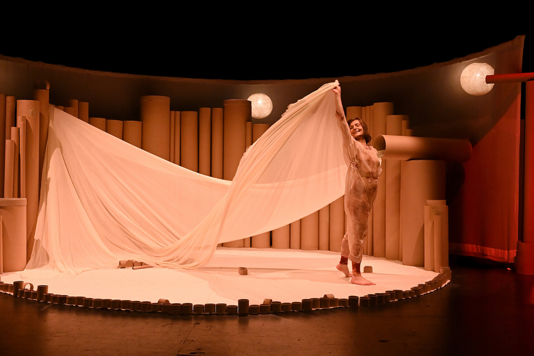 L'interprète sourit en tenant un grand voile translucide beige qui s'étend en éventail au-dessus d'elle. La scène est circulaire, bordée de petits anneaux de carton, avec une lune lumineuse projetée en arrière-plan.