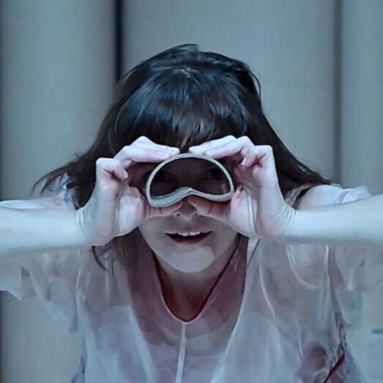 Le personnage de l'enfant regardant à travers deux tubes de carton formant des jumelles improvisées. Elle sourit légèrement et semble observer quelque chose avec curiosité dans une lumière bleutée.