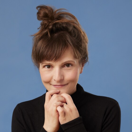 Portrait d’une femme souriante, vêtue d’un chandail noir à col roulé, posant devant un fond bleu uni. Elle a les cheveux bruns relevés en chignon désordonné et tient ses mains près de son visage d’un air doux et bienveillant.