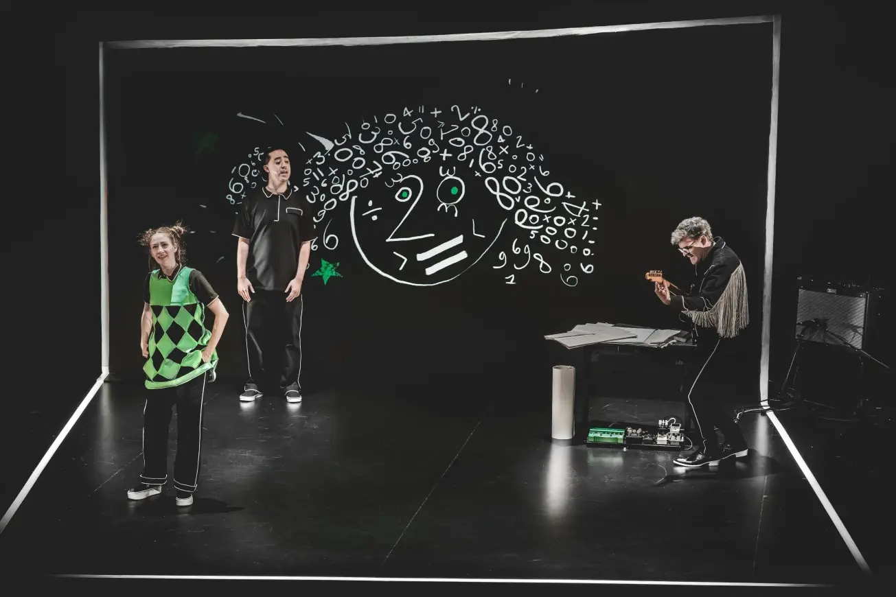 Trois personnages vêtus de vert et noir sont sur la scène, devant un grand tableau noir sur lequel est dessiné la tête d'un bonhomme fait de chiffres.