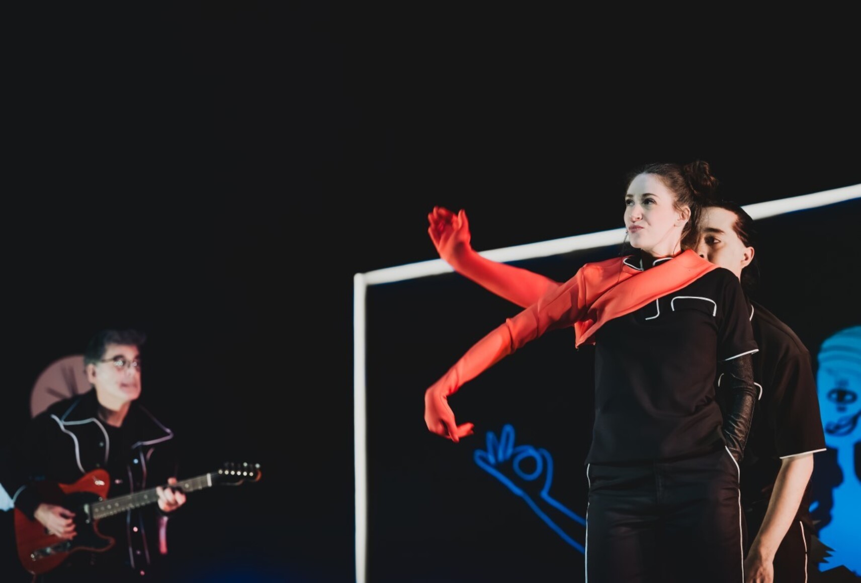 Un musicien joue de la guitare électrique. Deux personnages habillés en noir se tenant l'un derrière l'autre portent chacun un long gant rouge, créant une forme avec leurs bras. Ils sont devant un grand tableau noir avec de dessins bleus dessus.