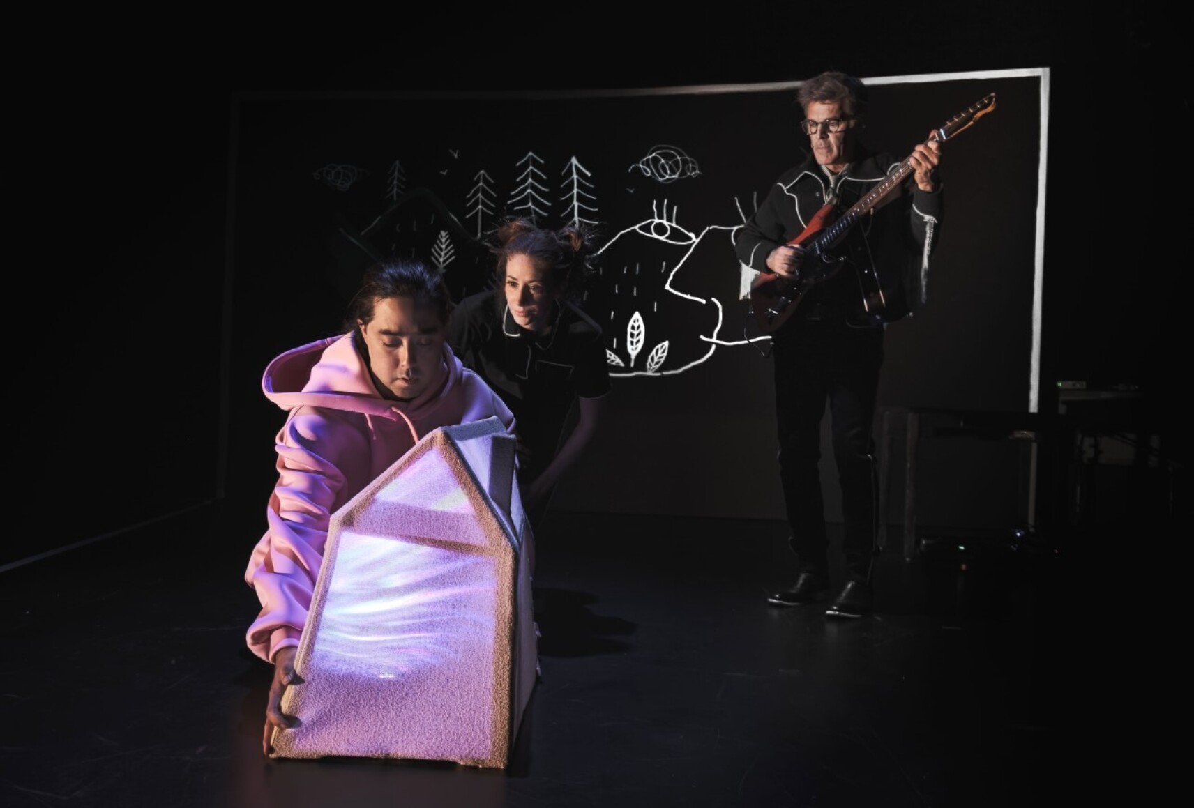 Un personnage porte un grand coton ouaté rose qui forme la structure d'une petite maison. Derrière lui, une comédienne habillée en noir et un musicien jouant de la guitare, devant un tableau noir avec un dessin de paysage tracé en blanc.