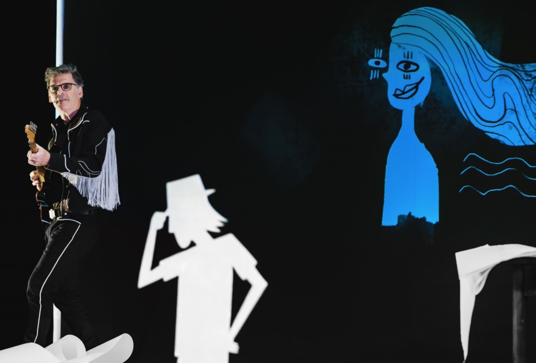 Photo d'un musicien jouant de la guitare. Derrière lui, sur un grand tableau noir, une silhouette blanche et un personnage féminin bleue.