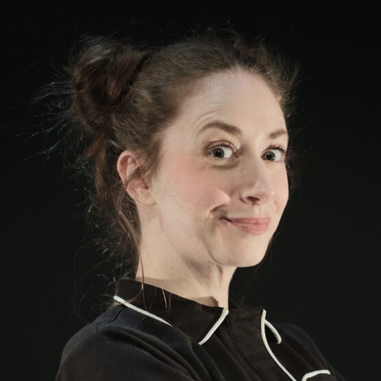 Photo du personnage de la jeune élève. Elle a les cheveux attachés en chignon et porte un costume noir aux contours blancs. Elle regarde est légèrement de profil, ses sourcils sont levés et elle regarde la caméra en souriant.