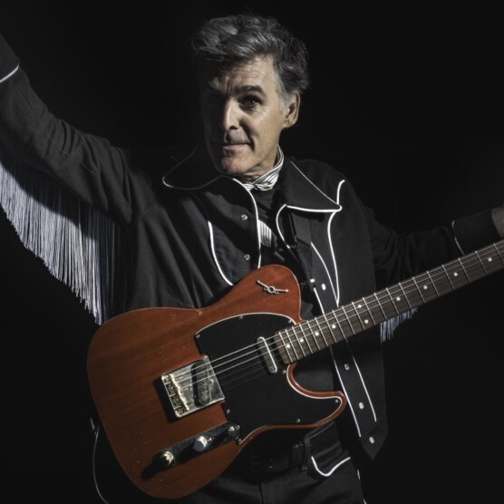 Monsieur Pierre porte une veste à franges noire, a une guitare électrique et lève le bras droit dans les airs en regardant la caméra.