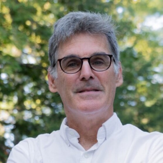 Photo de Pierre, un homme à la peau claire, aux cheveux gris courts, portant des lunettes aux montures noires et une chemise blanche. Il est dehors devant des arbres et regarde l'objectif.