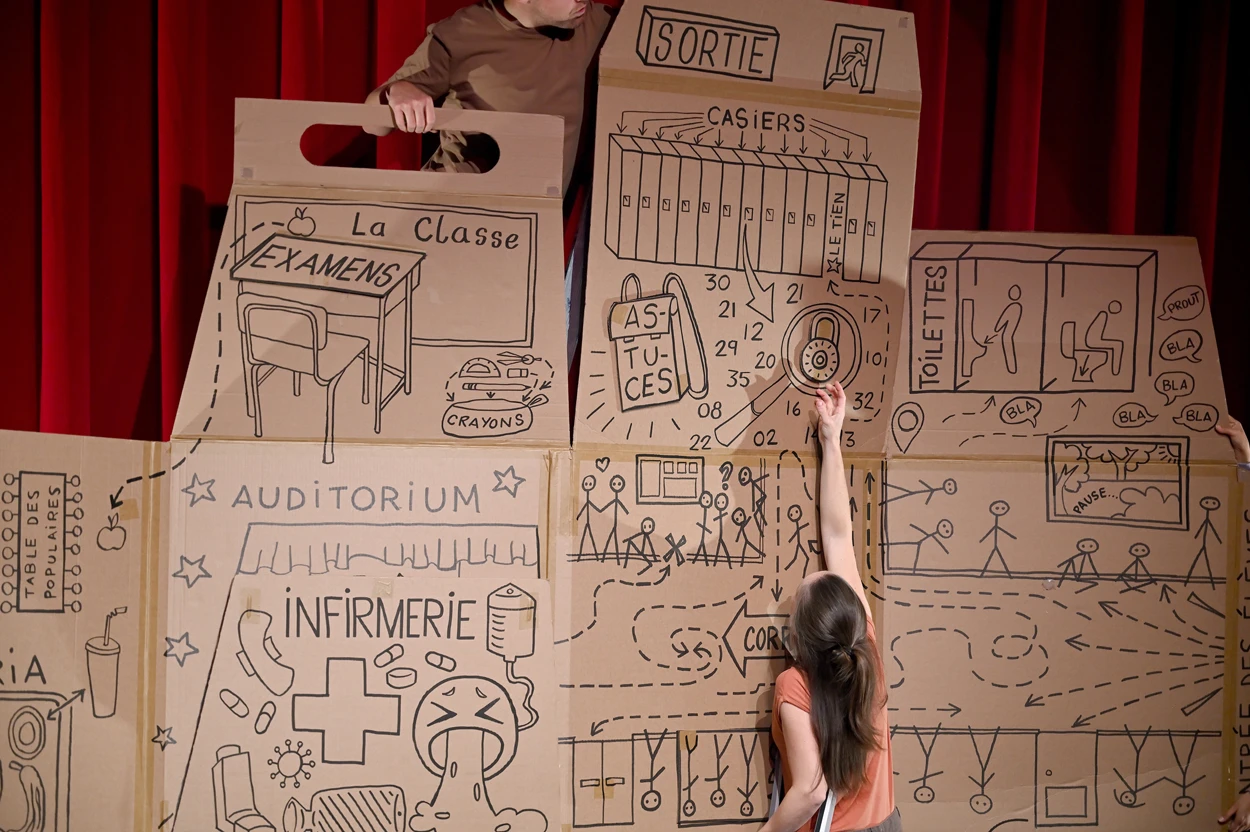 Une comédienne se tient de dos, bras levé, devant un immense carton qui prend presque toute la photo. Au fond, un rideau rouge. Sur le cartons, il y a de nombreux dessins et des mots : "classe" "examens" "auditorium" "infirmerie" "casiers" "astuces".