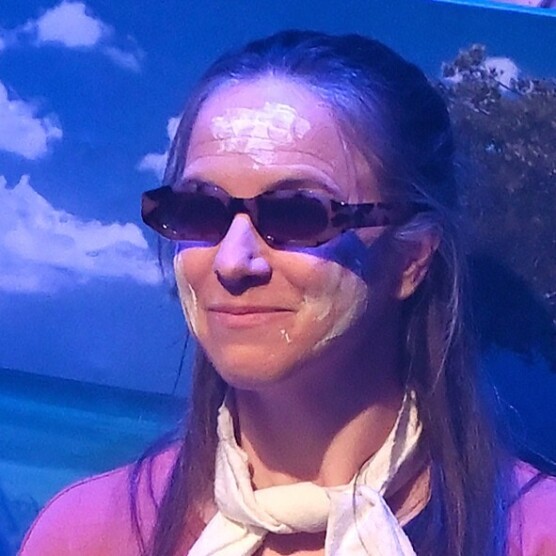 Plan sur le visage d'une interprète qui porte des lunettes de soleil, devant une image d'un ciel bleu et d'un océan. Elle a des traces blanches sur le visage et porte un foulard blanc noué autour du cou. Ses cheveux bruns tombent sur ses épaules.