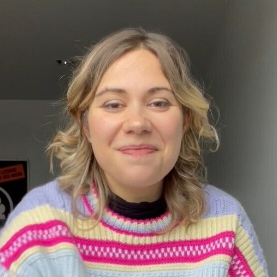 Une personne blonde aux cheveux mi-longs et bouclés regarde l'objectif en souriant. Elle porte un chandail à rayures rose, jaune et violet.