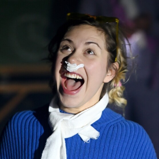 Plan sur le visage d'une personne blonde aux cheveux longs très expressive, qui semble crier en regardant vers le ciel. Elle a du coton dans le nez, porte un chandail bleu et un foulard blanc noué autour du cou.