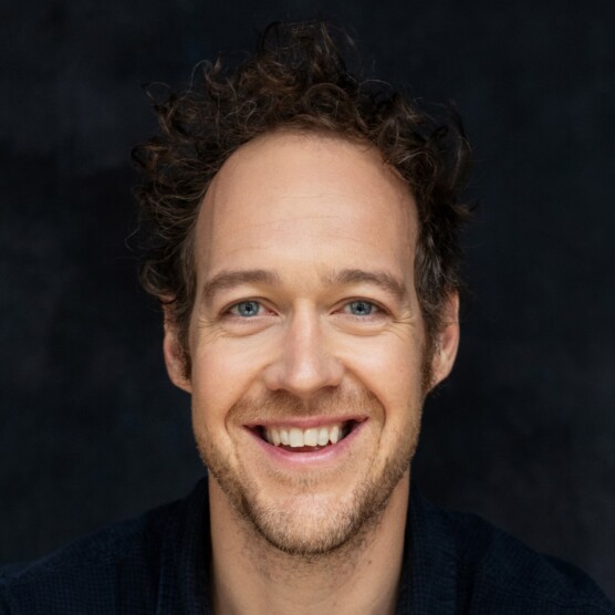 Portrait sur fond gris de David Leblanc, un homme aux cheveux châtains bouclés, aux yeux bleus et à la peau claire. Il a une barbe de quelques jours et sourit largement.