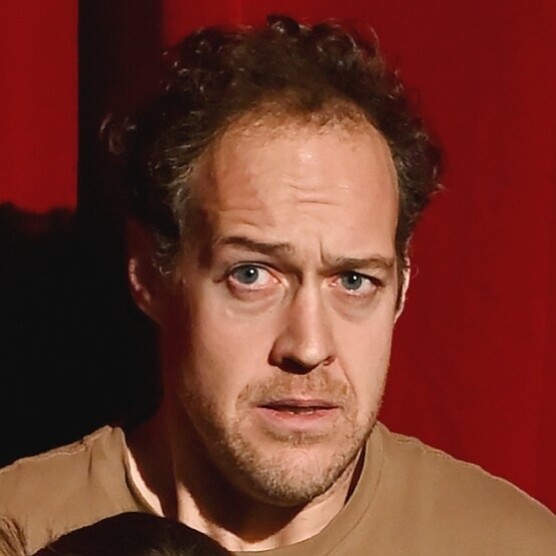 Gros plan sur le visage de David, sur fond rouge. Il a le visage tendu, le regard inquiet et semble préoccupé.