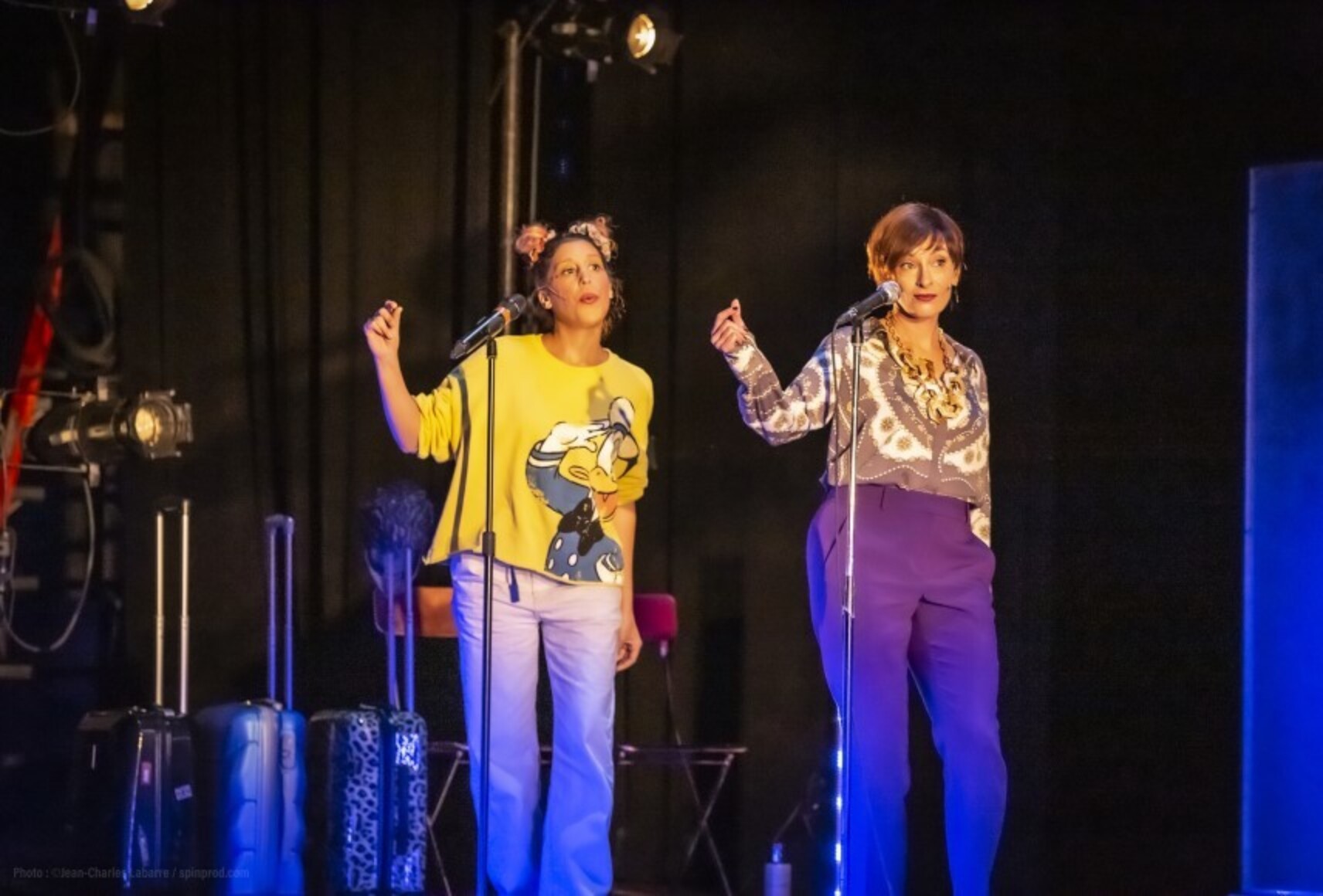 Angela et Jade chantent et claquent des doigts devant un micro. Derrière elles, trois valises et des chaises alignées.