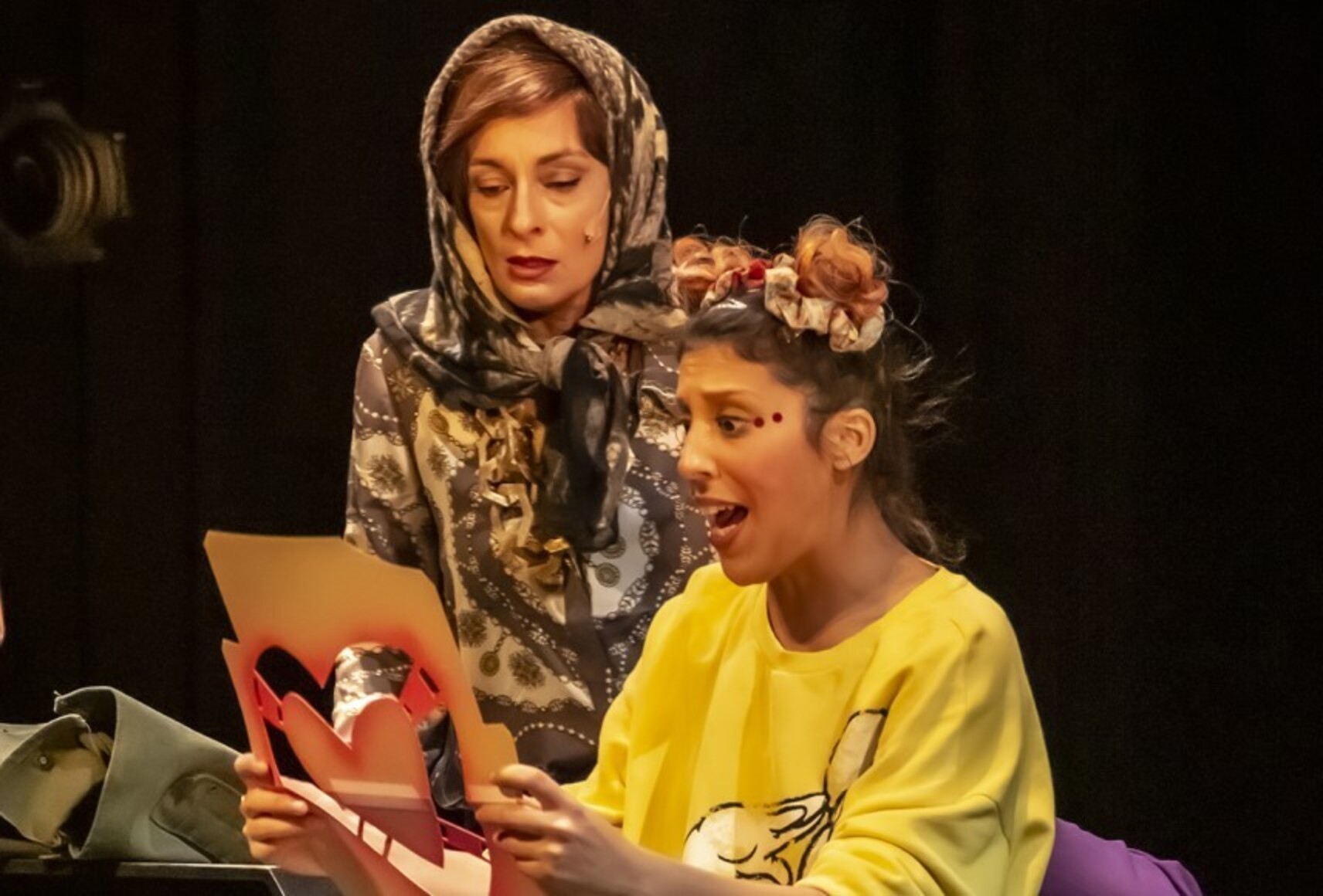 Jade étonnée devant un pochoir, Angela la regarde. Jade porte un chandail jaune et deux chignons sur sa tête. Angela porte une blouse et un foulard à motifs sur sa tête.