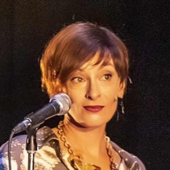 Photo d'Angela, qui porte les cheveux courts détachés, du rouge à lèvres foncé et une chemise à motifs. Un micro sur pied à sa droite est dirigé vers elle et son regard est dirigé vers sa gauche.