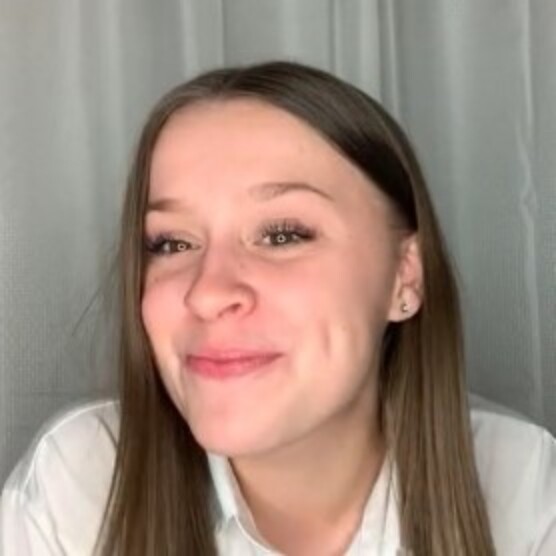 Photo de Madison, une femme au teint clair, aux longs cheveux bruns lisses et aux yeux bruns. Son visage est légèrement tourné vers la gauche et elle regarde devant elle en souriant. Elle porte un haut blanc et est posée devant un fond gris.