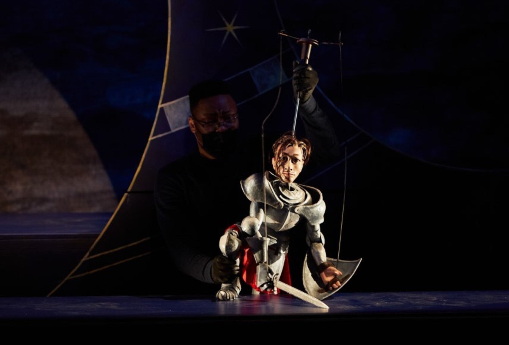 Le personnage Orlando, seul en scène, sans son casque d'armure, un genou au sol et son arme déposée. Il est éclairé par une lumière blanche et le reste de la scène et du décor sont dans l'obscurité.