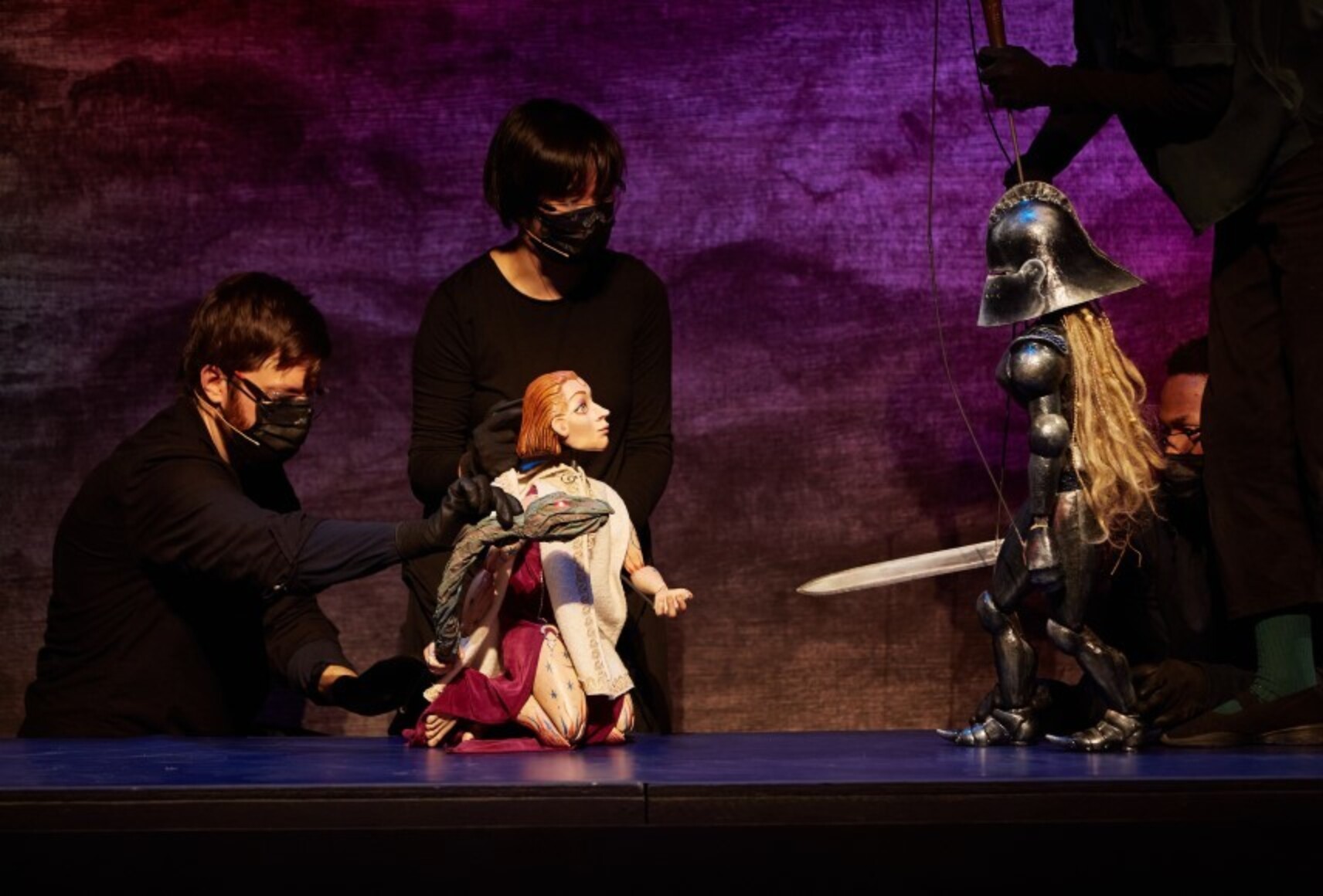 Des acteurs habillés en noir manipulent des marionnettes dans la lumière. Mélisse (la magicienne) à genoux, et Bradamante (la chevalière du Nord) se font face, devant un décor aux couleurs mauves.